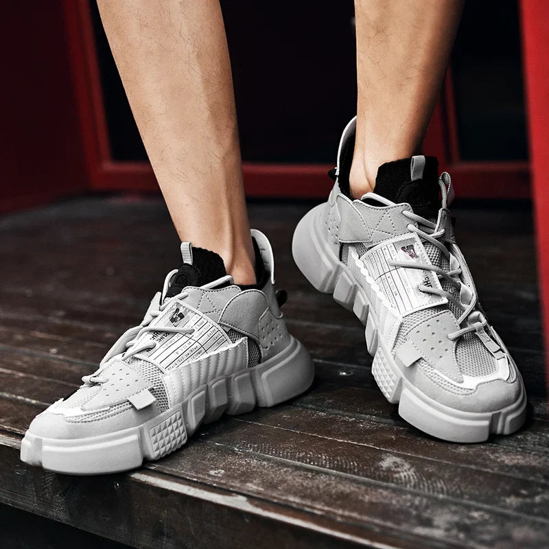 A person wearing chunky gray Extreme VS7 Sneakers with thick, textured soles stands on a dark wooden floor, their legs visible from the knee down. These stylish streetwear sneakers feature mesh and leather details.