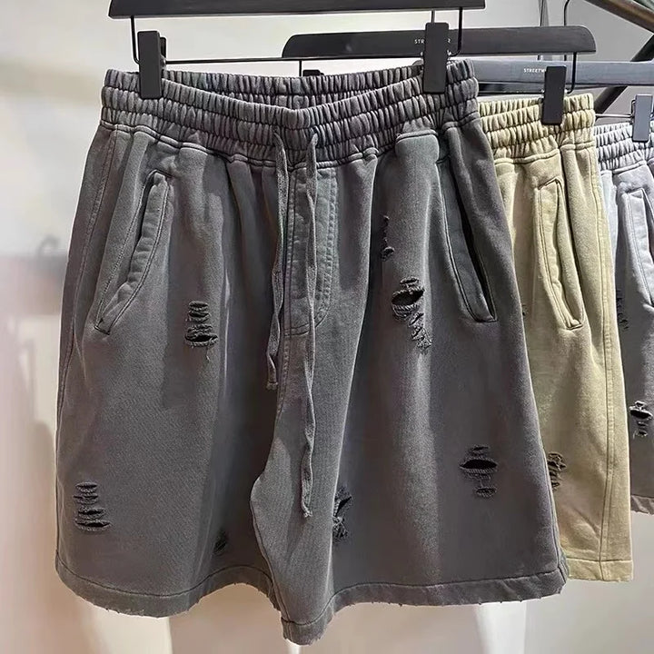 Gray distressed shorts with an elastic waistband and drawstring, showcasing a cut-up design with multiple rips and frayed patches. Another similar pair of vintage baggy shorts in beige hangs beside them on black hangers.