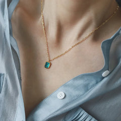 A person wearing a light blue, partially unbuttoned shirt and a hypoallergenic gold chain necklace with a small, rectangular turquoise pendant. The focus is on the necklace and collarbone area.