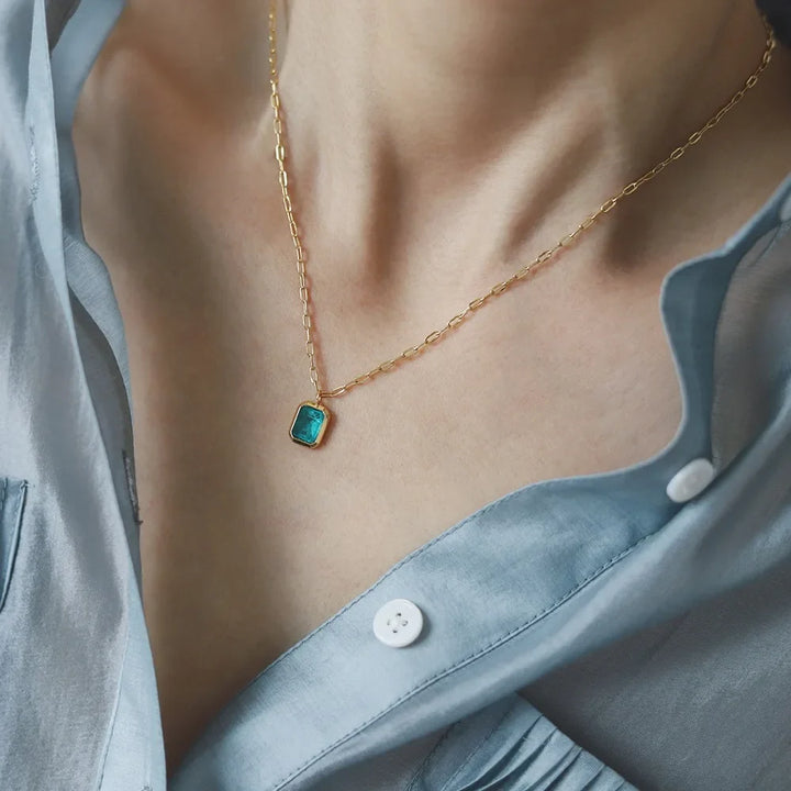 A person wearing a light blue, partially unbuttoned shirt and a hypoallergenic gold chain necklace with a small, rectangular turquoise pendant. The focus is on the necklace and collarbone area.