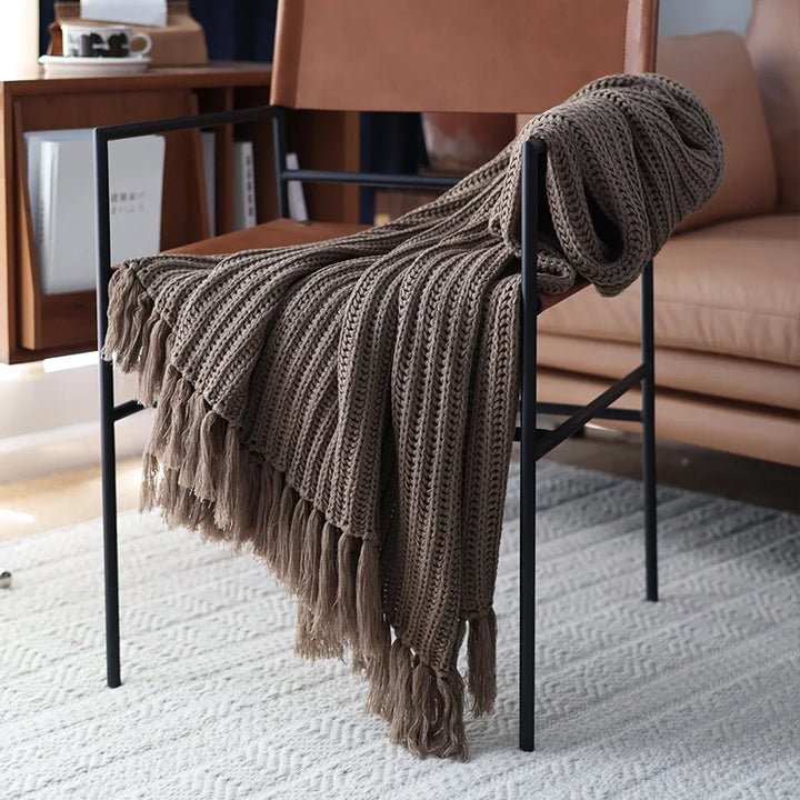 A brown, earth-tone throw with tassels drapes over the arm and seat of a modern brown leather chair with black metal legs, set on a textured light-colored rug.