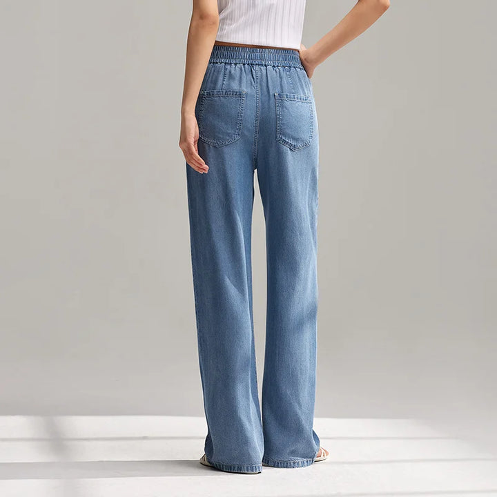 A person wearing light blue, wide-leg denim pants with an elastic waistband and back pockets stands barefoot, facing away from the camera against a neutral background.