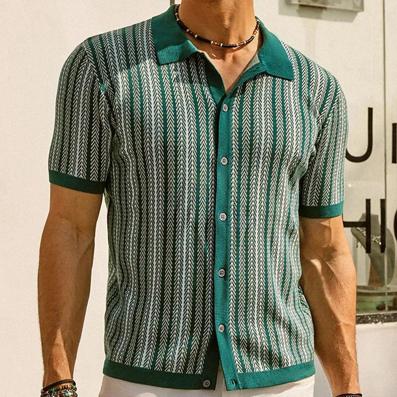 A man wearing a green short-sleeve Italian linen polo with a white chevron pattern stands indoors. He has a necklace and bracelets on, and part of a sign is visible in the background.