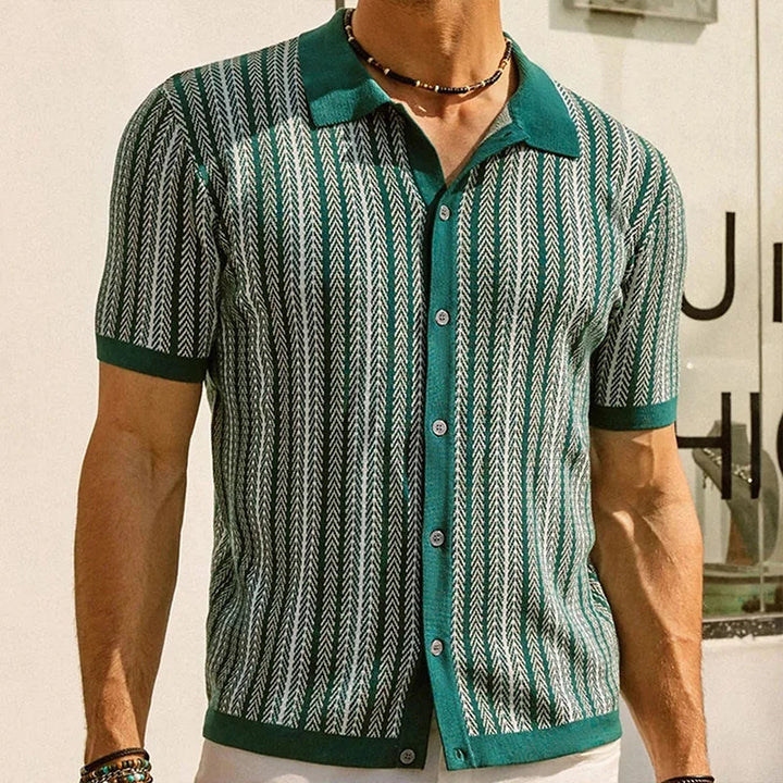 A man wearing a green short-sleeve Italian linen polo with a white chevron pattern stands indoors. He has a necklace and bracelets on, and part of a sign is visible in the background.