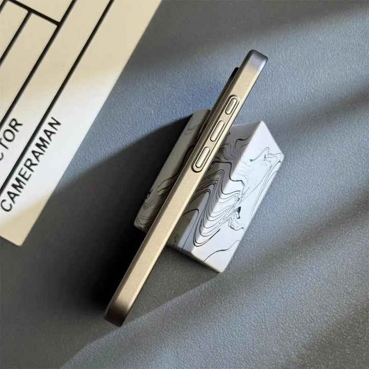 A smartphone with a slim lightweight design and metallic edges rests upright on a marble-patterned stand, positioned on a gray surface. Part of a clapperboard with the word “CAMERAMAN” is visible in the upper left corner.