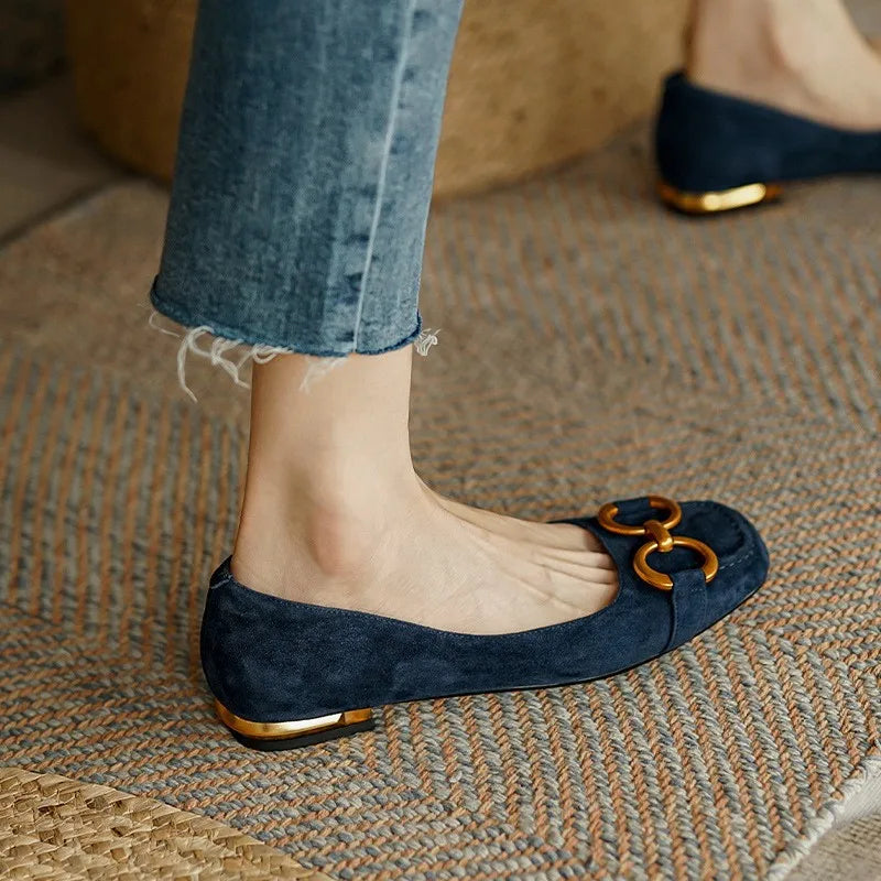 A person wearing blue suede ballet flats with gold decorative buckles and metallic accents on the heels, paired with frayed hem jeans, stands on a textured woven rug.