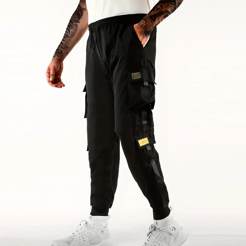 A person wearing black cargo pants with side pockets, ribbed ankle cuffs, and an elastic waist, paired with white sneakers and tattooed arms, stands against a plain light background.