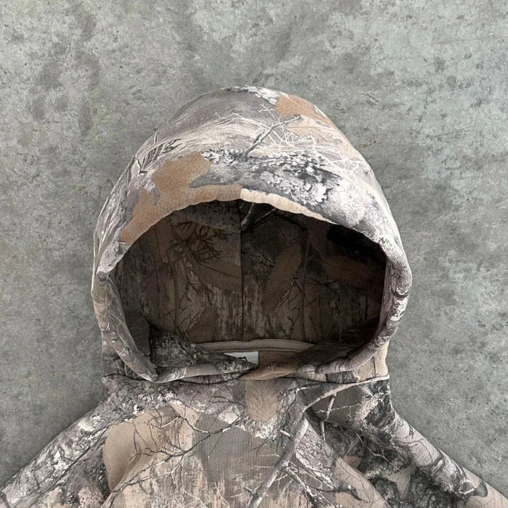 A hooded jacket with an oversized fit and gray-brown camo pattern is laid flat on a stone-like gray surface, with the hood facing upward.