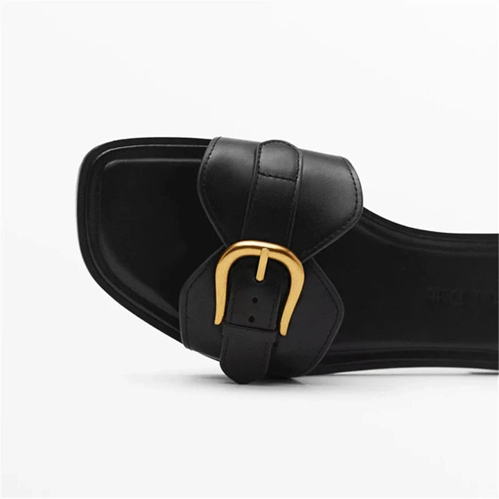 A close-up of a black leather slide sandal with a square toe and a gold buckle detail on the strap, set against a plain white background.