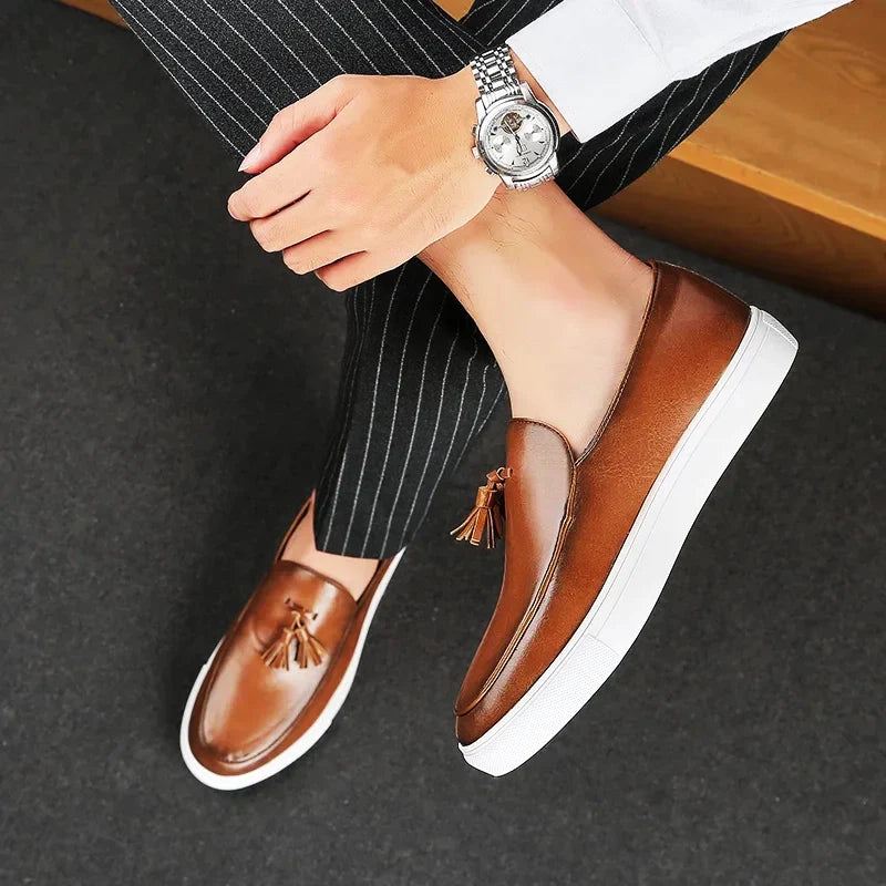 A person wearing brown luxury leather loafers with white soles, pinstripe pants, a white shirt, and a silver wristwatch sits with legs crossed on a dark carpeted floor. The cushioned insole adds comfort to their stylish look.