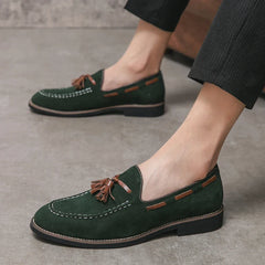 A person wearing dark green tassel loafers with brown trim and a cushioned footbed, paired with black pinstripe pants, stands on a gray concrete floor.