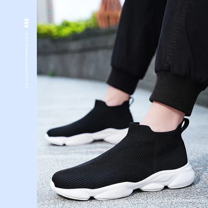 A person wearing lightweight black slip-on sneakers with a non-slip sole and thick white base stands on a paved surface outdoors. Only their lower legs and feet are visible, dressed in black pants.