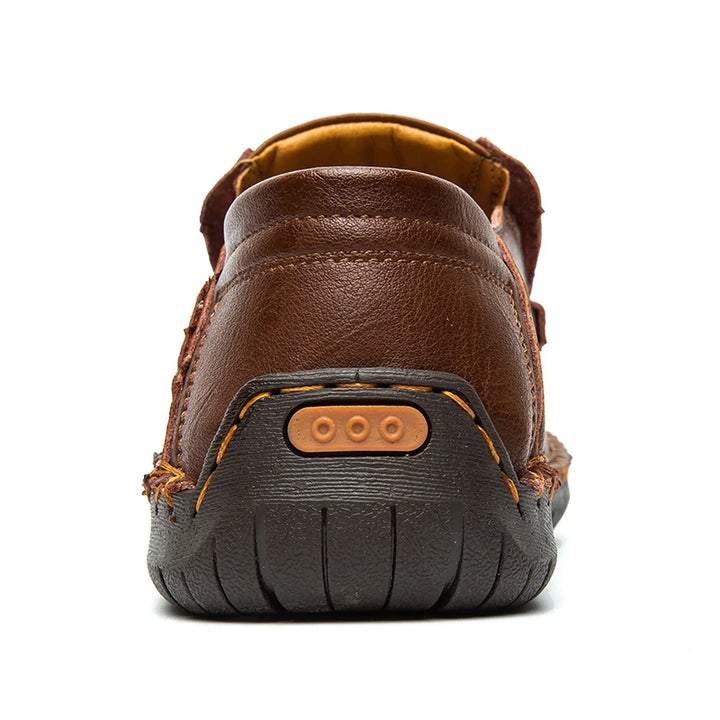 A close-up rear view of a brown leather loafer showcases traditional craftsmanship with detailed stitching, a textured black rubber sole, and a small tan accent on the heel.