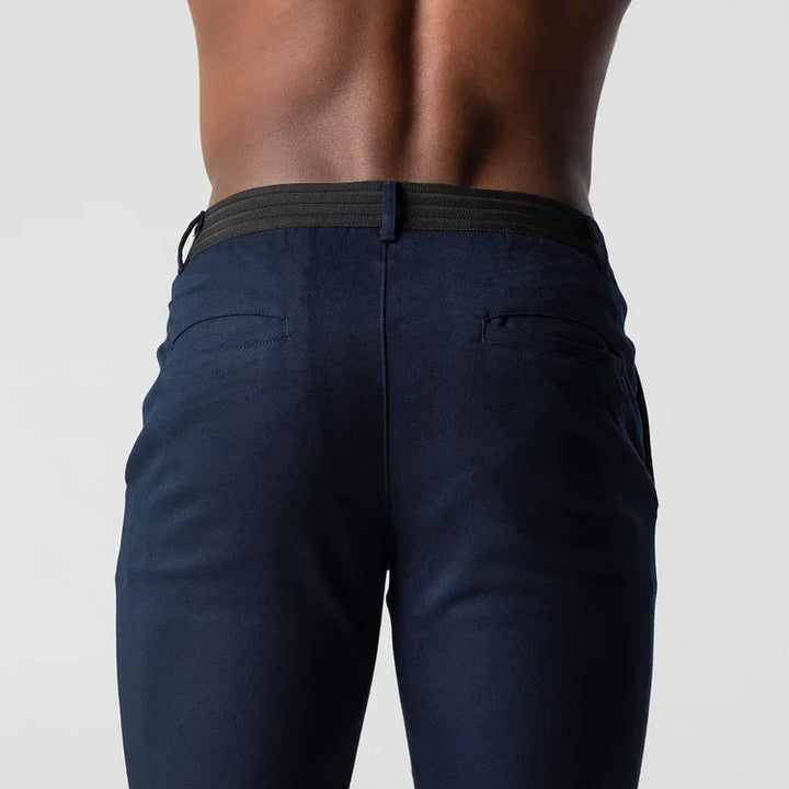 A close-up back view of a person wearing dark blue, slim fit pants with a black waistband, showing the lower back and upper legs against a plain light background.