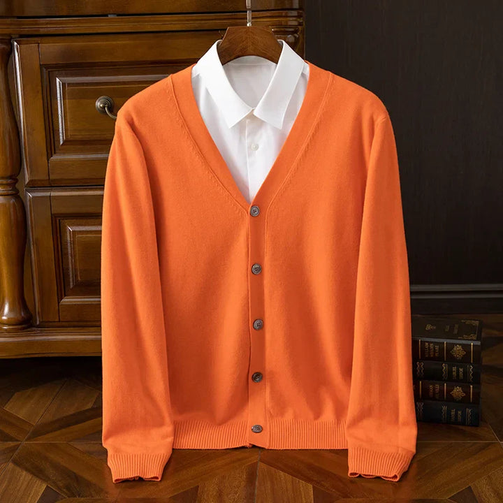 An orange button-up cardigan with a relaxed fit and V-neckline is layered over a white collared shirt, hanging on a wooden hanger in front of a dresser, with books stacked nearby on the wood floor.