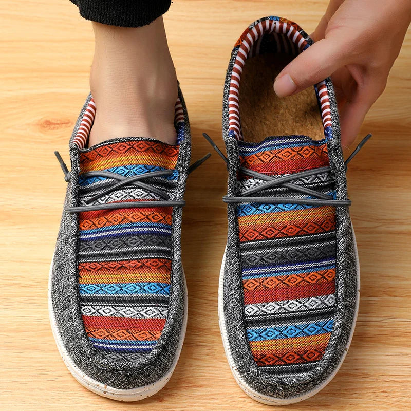 A person wears colorful slip-on shoes with a vibrant ethnic pattern of geometric designs in orange, blue, and black on a light wooden floor. One woven upper shoe is being adjusted by hand.