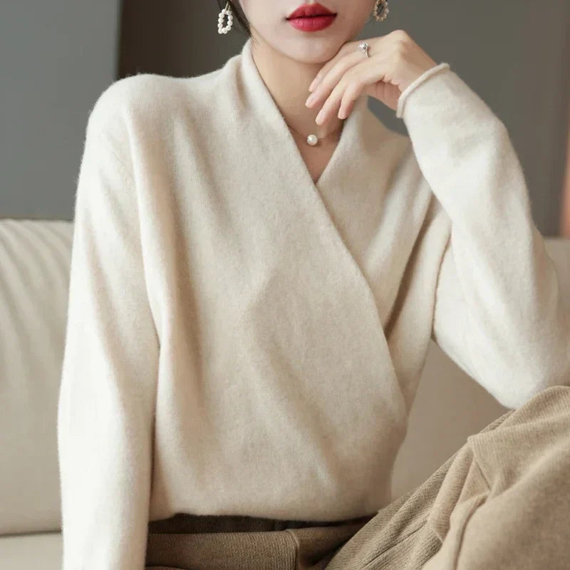 A person wearing a soft, cream-colored cashmere wrap sweater and light brown pants sits on a beige couch, resting their chin on their hand. They have short hair, red lipstick, and are accessorized with earrings and a ring.