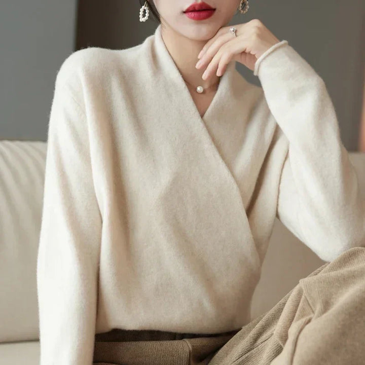 A person wearing a soft, cream-colored cashmere wrap sweater and light brown pants sits on a beige couch, resting their chin on their hand. They have short hair, red lipstick, and are accessorized with earrings and a ring.