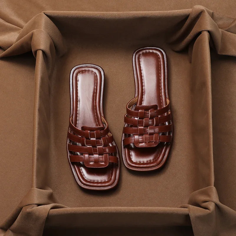 A pair of brown, woven Italian leather slip-on sandals with a square-toe design, displayed on a soft brown fabric background and framed by draped cloth.