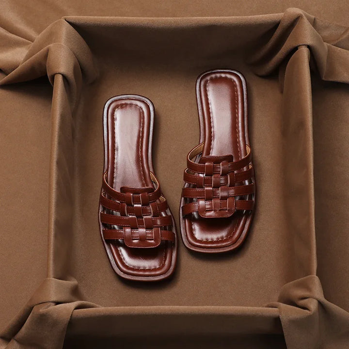 A pair of brown, woven Italian leather slip-on sandals with a square-toe design, displayed on a soft brown fabric background and framed by draped cloth.