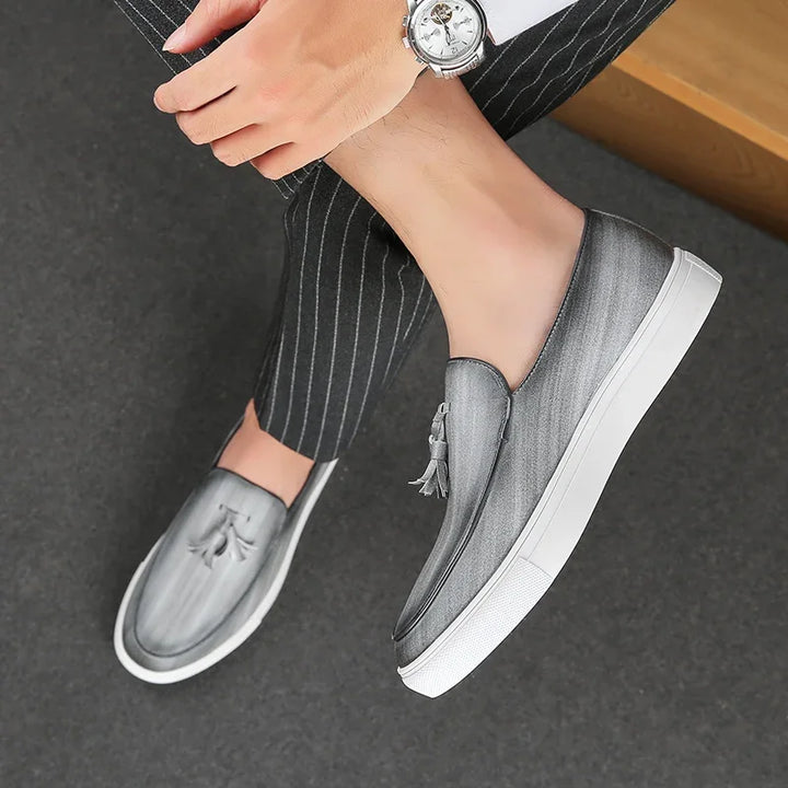 A person wearing grey tassel loafers with cushioned insoles and white soles sits with legs crossed, dressed in pinstriped pants, and wearing a silver wristwatch.