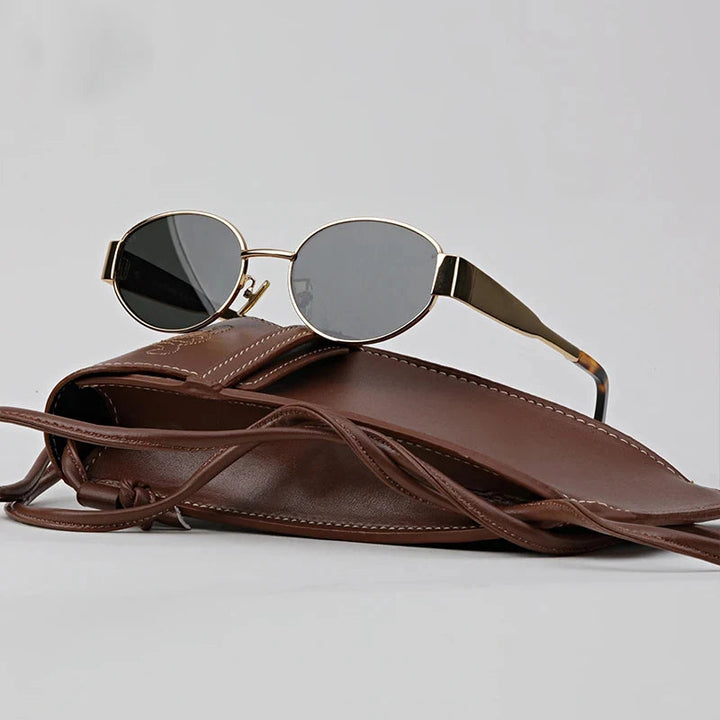 Round, gold-framed vintage-inspired sunglasses with dark lenses rest on top of a brown leather case with a strap, against a plain gray background.