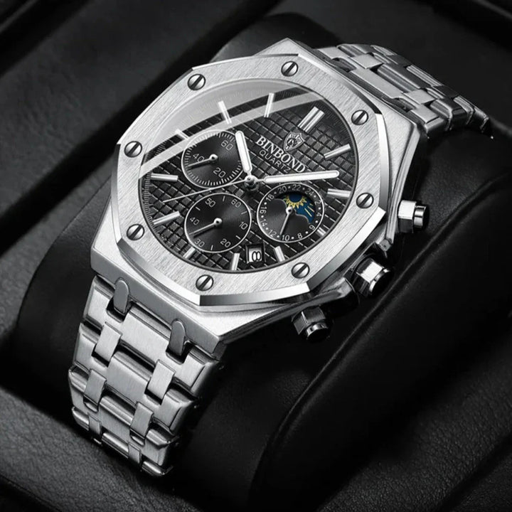 A luxury stainless steel watch with a textured black dial, three chronograph dials, and a metal link band, displayed on a black leather cushion. The BINBOND face highlights a moon phase indicator for added elegance.