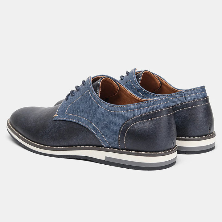 A pair of comfortable oxfords in dark blue leather with denim fabric accents, white stitching, cushioned sole, brown inner lining, and white and black rubber soles, displayed on a light background.