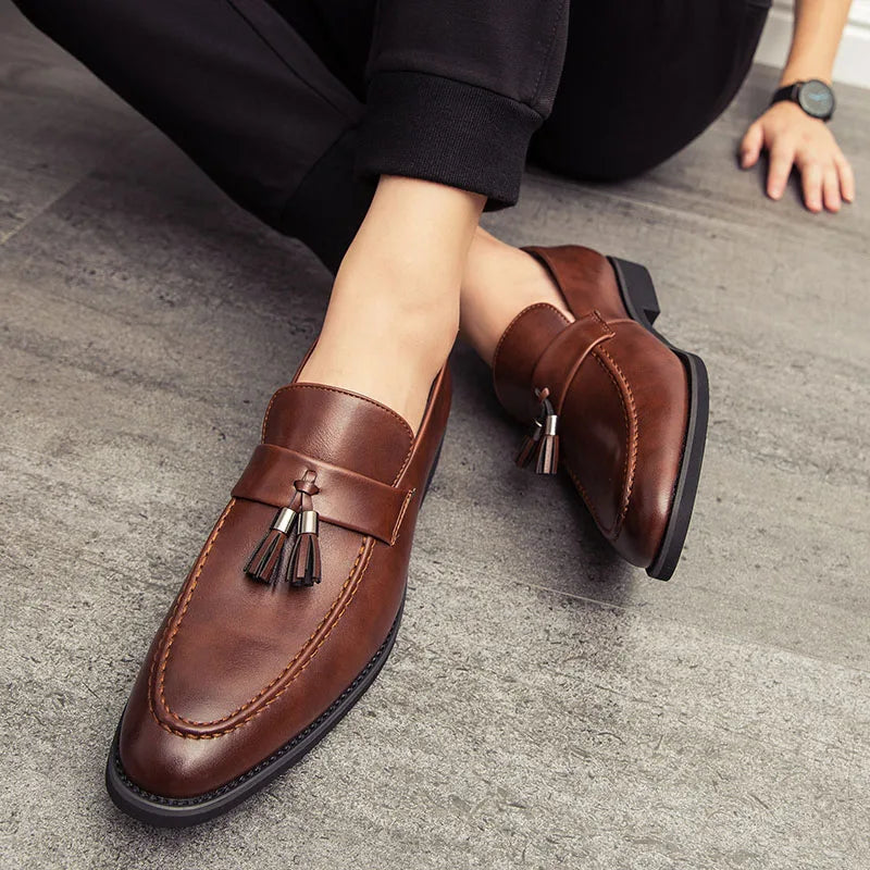 A person sits on the floor wearing polished leather tassel loafers with black soles. The minimalistic shoes are paired with black pants for a refined look.