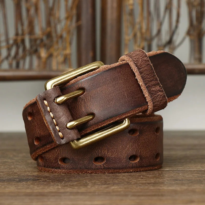A rolled-up genuine leather belt with a copper buckle and stitched edges sits on a wooden surface, set against a rustic background. The belt showcases heritage craftsmanship, featuring multiple holes along its length.