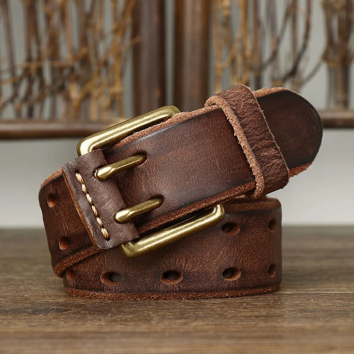 A rolled-up genuine leather belt with a copper buckle and stitched edges sits on a wooden surface, set against a rustic background. The belt showcases heritage craftsmanship, featuring multiple holes along its length.