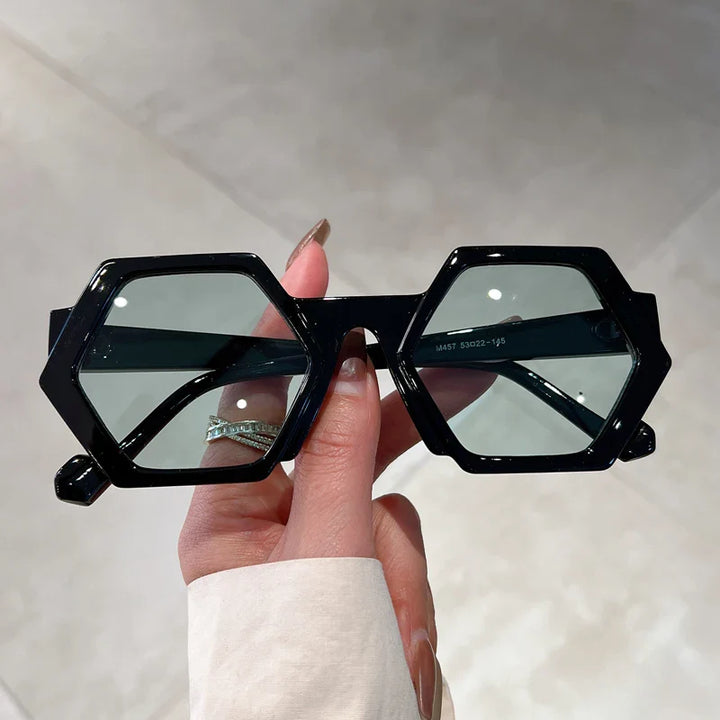 A hand with manicured nails holds a pair of trend-forward, hexagonal frame sunglasses featuring tinted lenses and UV400 protection against a light, blurred background.