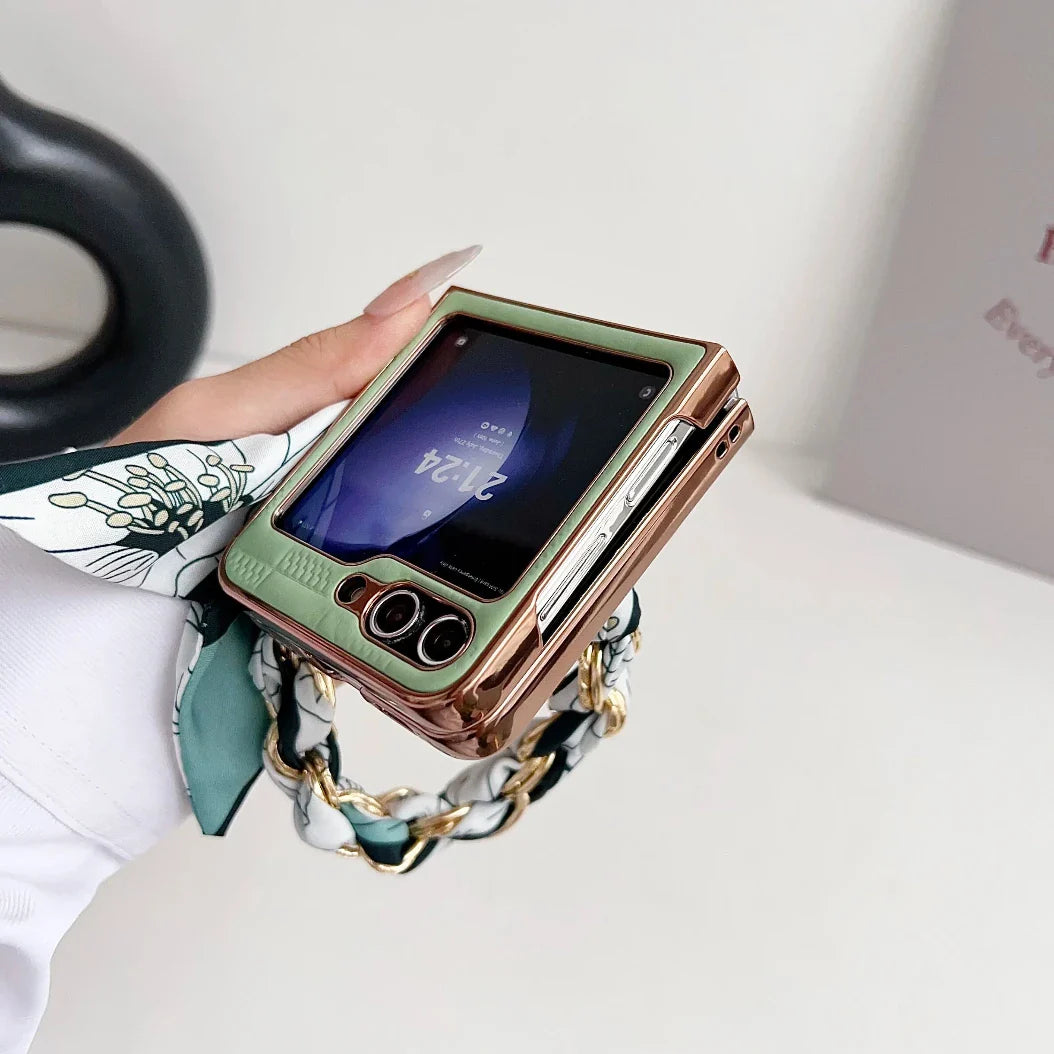A hand holding a Samsung Galaxy Z Flip with a green and gold premium leather case, displaying the time on its small external screen. The person has manicured nails and is wearing a patterned scarf wrapped around the wrist.