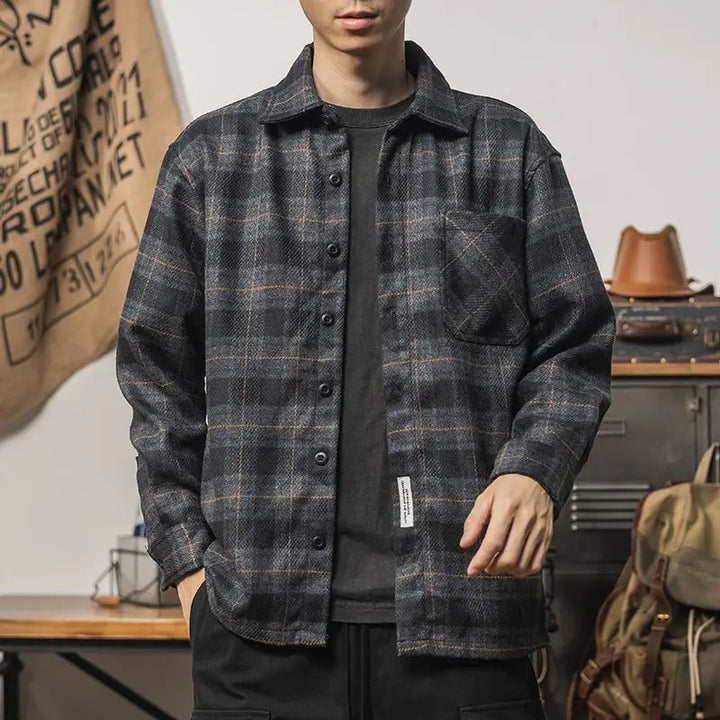 A person wears a relaxed fit black and gray plaid button-up shirt over a black t-shirt, standing indoors with one hand in their pocket. The background includes a desk, bags, and a tan wall hanging with text.