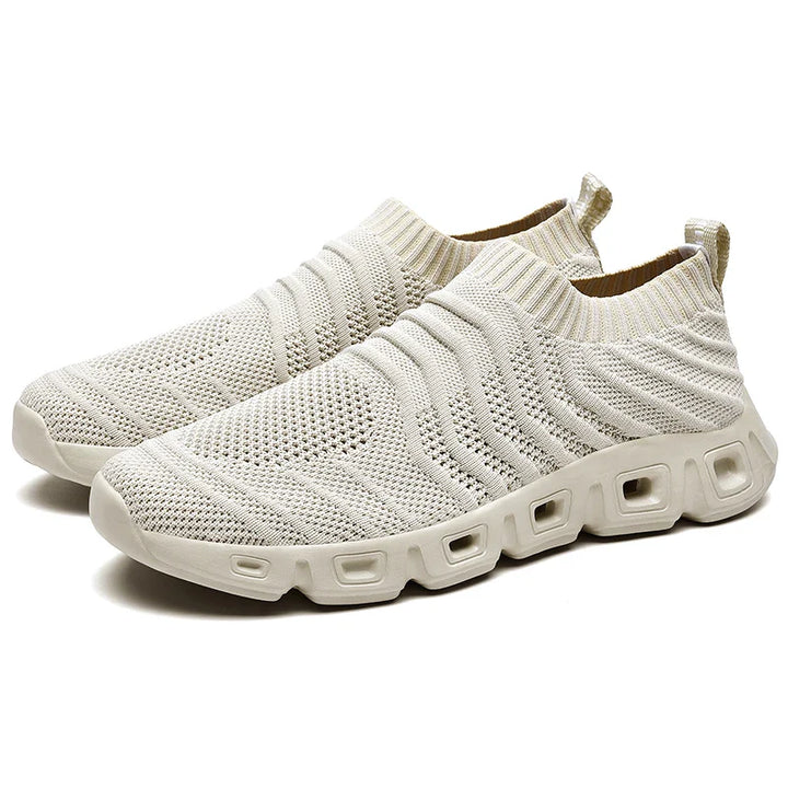 A pair of beige slip-on athletic sneakers with a textured, moisture-wicking knitted upper and a chunky, cushioned sole featuring rectangular cutouts for added comfort and style.