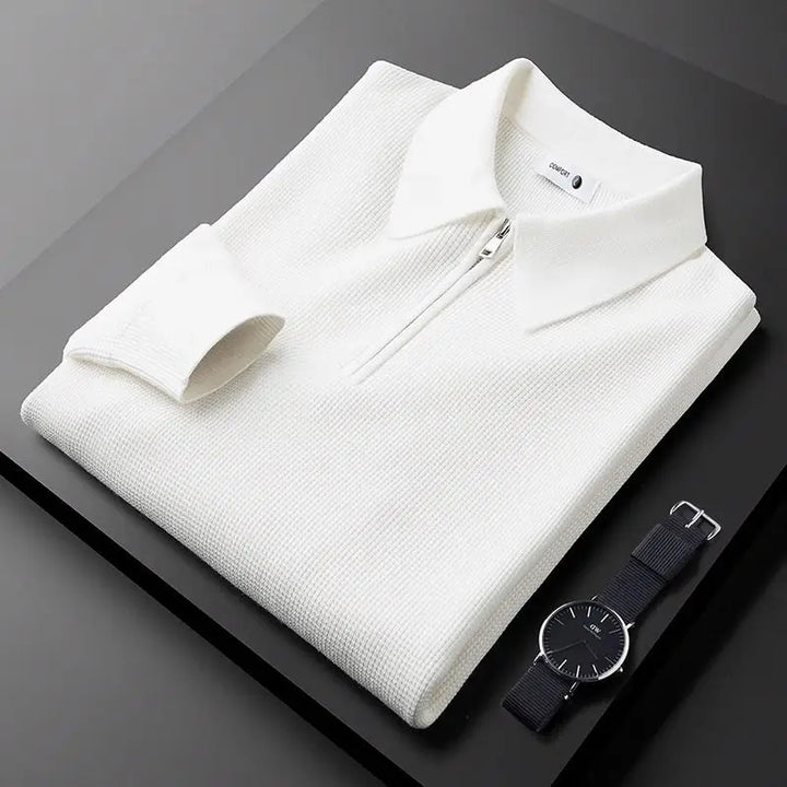 A neatly folded white textured long sleeve polo with a collar is placed on a black surface next to a black wristwatch with a dark strap and a minimalist face.