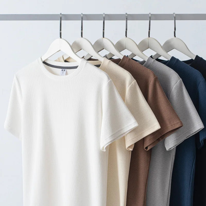 Six short-sleeved t-shirts in white, beige, light brown, brown, gray, and navy—crafted from soft 260G cotton—hang neatly on white hangers from a metal rail against a plain white background.