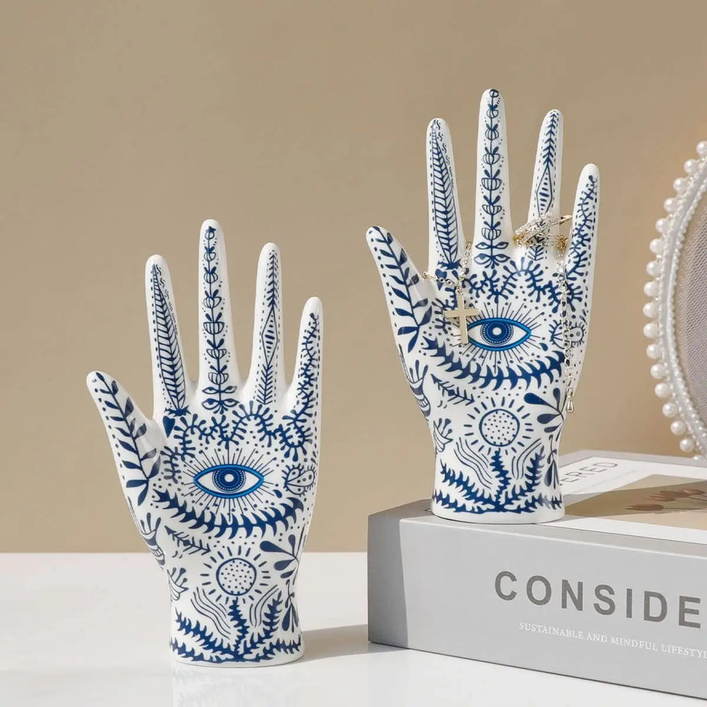 Two white ceramic hand sculptures with blue botanical patterns and eye motifs evoke boho-chic home décor. One ceramic jewelry stand rests on a box labeled CONSID, while the other stands beside it, enhancing your evil eye decor.