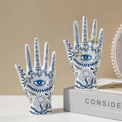 Two white ceramic hand sculptures with blue botanical patterns and eye motifs evoke boho-chic home décor. One ceramic jewelry stand rests on a box labeled CONSID, while the other stands beside it, enhancing your evil eye decor.