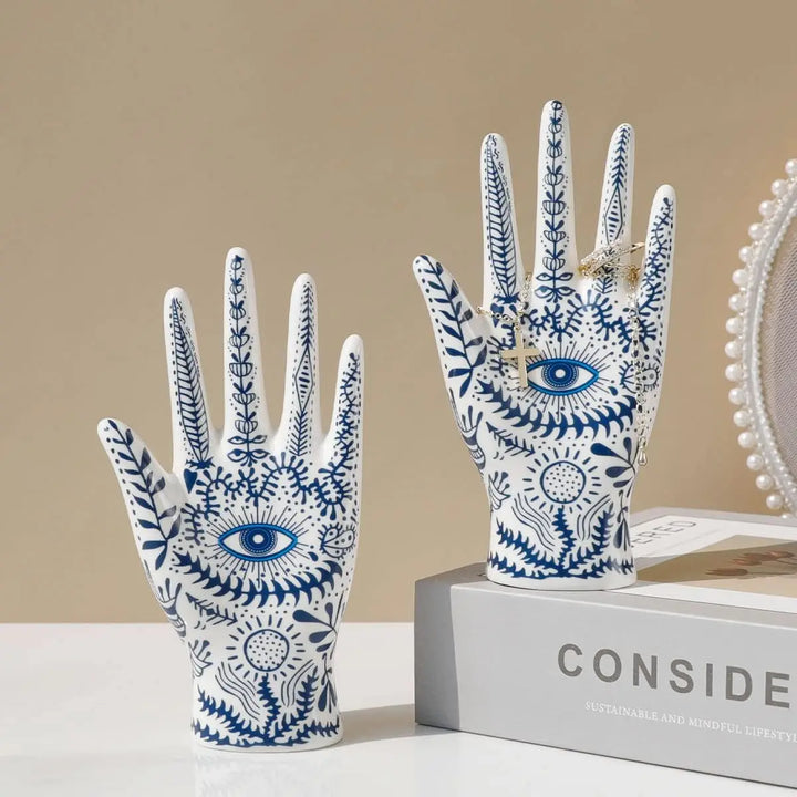 Two white ceramic hand sculptures with blue botanical patterns and eye motifs evoke boho-chic home décor. One ceramic jewelry stand rests on a box labeled CONSID, while the other stands beside it, enhancing your evil eye decor.