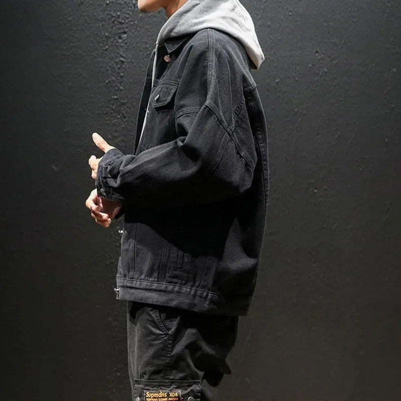 A person stands sideways against a dark background, wearing a black premium denim jacket over a gray hoodie and black pants with a visible patch near the pocket. The relaxed silhouette is complemented by their head slightly turned away.