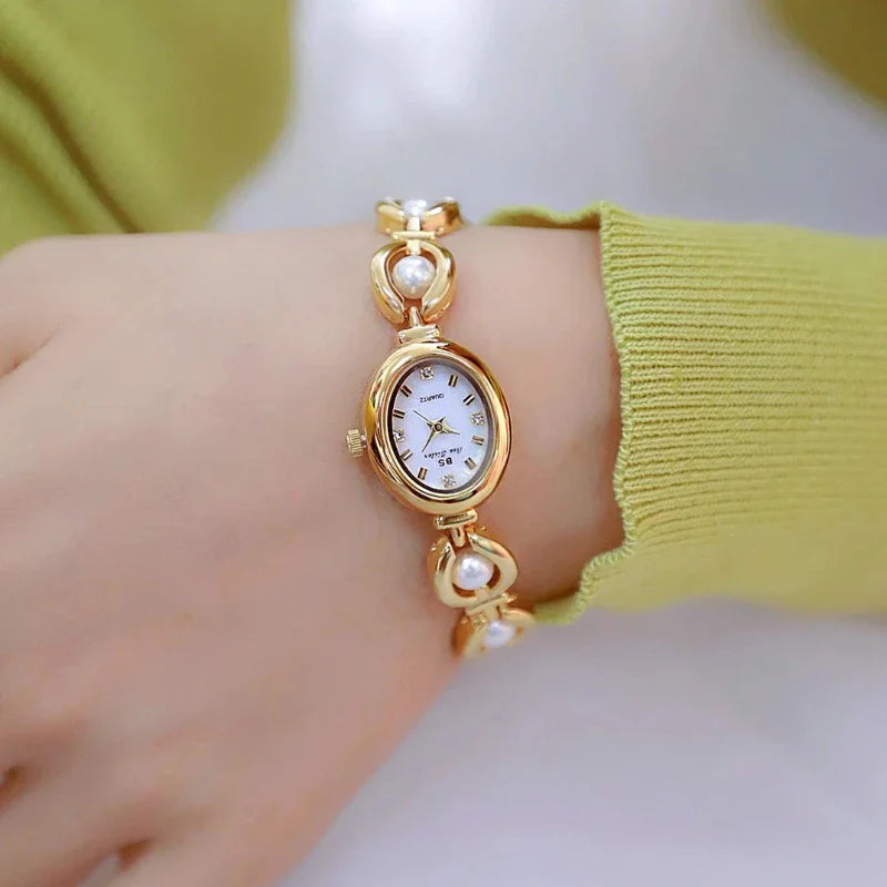 A gold wristwatch with a white oval face and Roman numerals, set on a bracelet adorned with pearl accents, features precision quartz movement and is worn on a wrist with a light green long-sleeve top.
