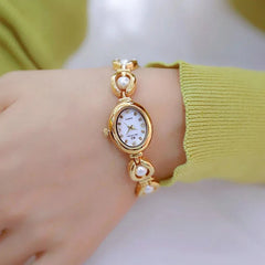 A gold wristwatch with a white oval face and Roman numerals, set on a bracelet adorned with pearl accents, features precision quartz movement and is worn on a wrist with a light green long-sleeve top.
