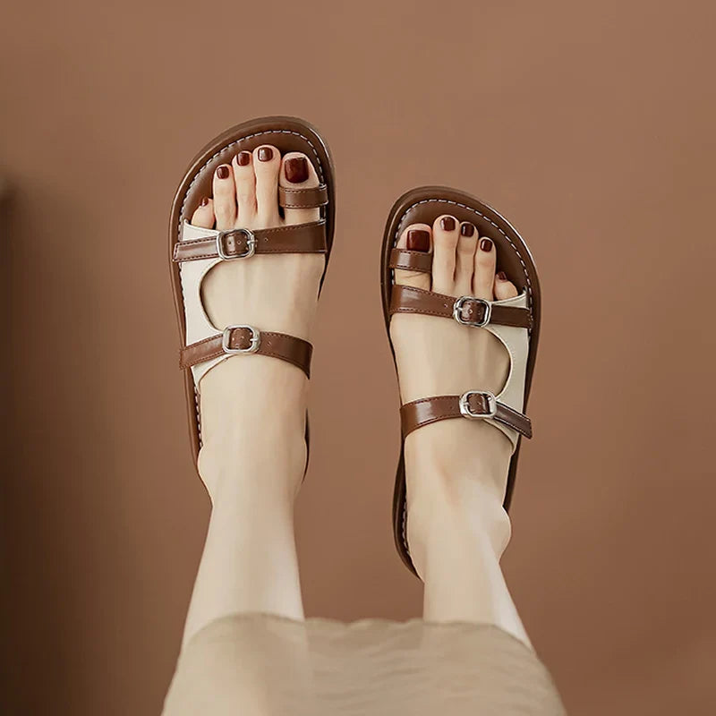 A person with light skin wears brown sandals featuring a leather upper and dual buckle straps, shown against a solid brown background. Their toenails are painted dark red.