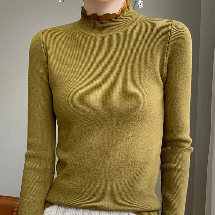 A person wearing a fitted, olive green ribbed sweater crafted from a cashmere blend with a high ruffled neckline and long sleeves, standing indoors against a neutral background.