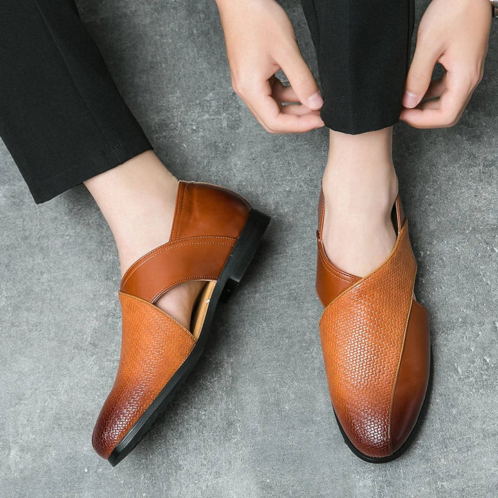A person wearing black pants is putting on stylish genuine leather loafers with a textured pattern and durable outsole, sitting on a gray textured floor.
