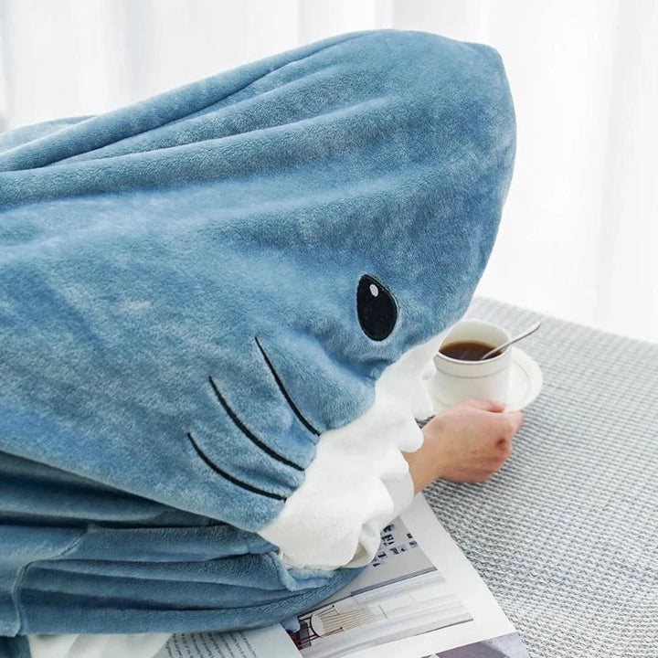 A person wrapped in blue shark pajamas sits at a table, holding a cup of tea or coffee. The cozy pajamas have a fin and eye, resembling a sharks head covering the persons upper body.
