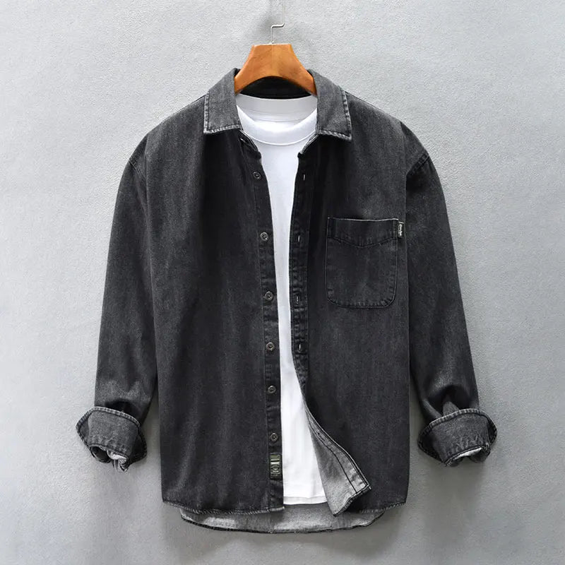 A dark grey washed denim overshirt with rolled sleeves hangs open over a plain white T-shirt on a wooden hanger against a light grey wall.