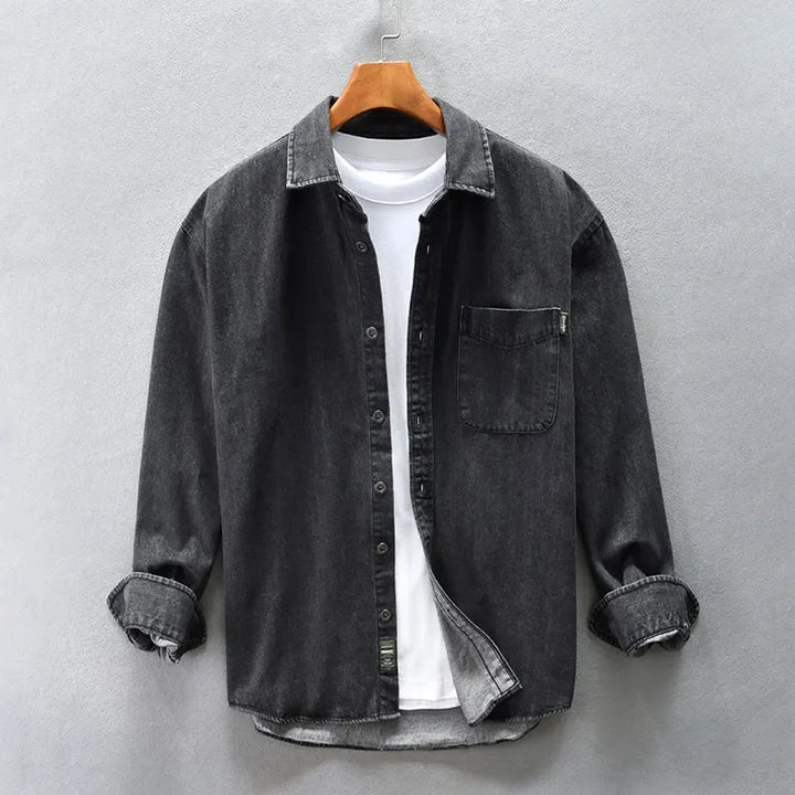 A dark grey washed denim overshirt with rolled sleeves hangs open over a plain white T-shirt on a wooden hanger against a light grey wall.