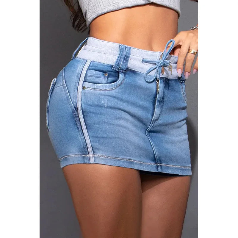 A woman wears a high waist denim skirt in light blue premium cotton, featuring front pockets, zipper details, and a tie at the waist. She pairs it with a cropped gray top, and her hand adorned with rings is visible.